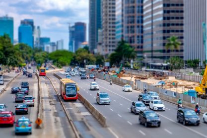 Photo of red public transport bus and many other vehicle, crossing primary road in Jakarta, Indonesia, captured in Jend. Sudirman street, with minature or tilt-shift effect.