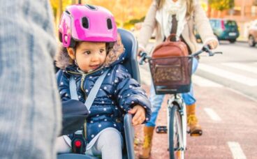 Portrait of little girl with security helmet on the head sitting in bike seat and her mother with bicycle on the background. Safe and child protection concept.