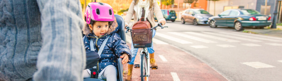 Portrait of little girl with security helmet on the head sitting in bike seat and her mother with bicycle on the background. Safe and child protection concept.