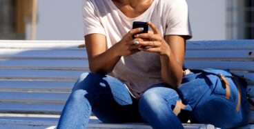 Portrait of beautiful young african woman sitting outdoors on bench with mobile phone and laughing