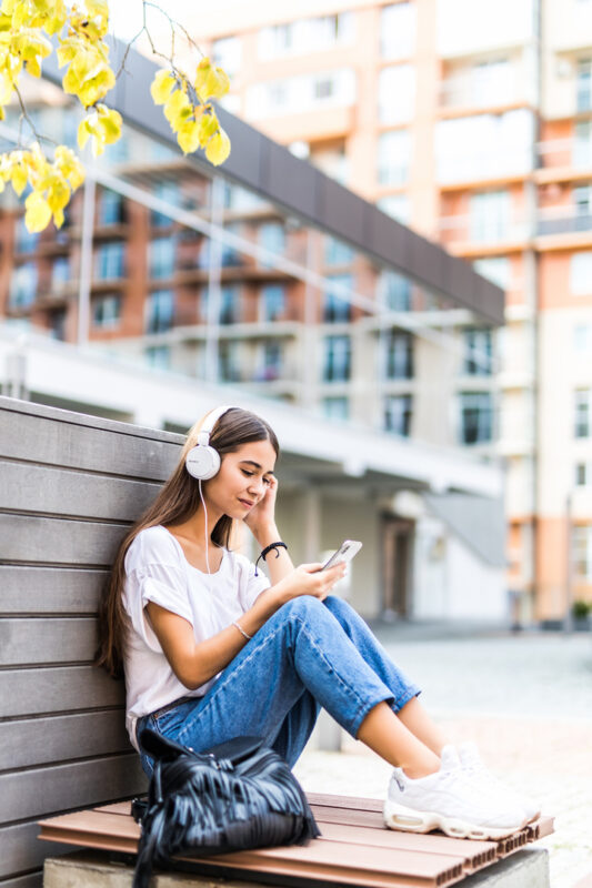 Portrait of smiling girl with earphones listening to music sitting on the bench outdoors