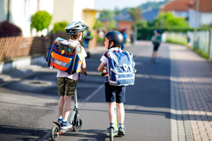 Two school kid boys in safety helmet riding with scooter in the city with backpack on sunny day. Happy children in colorful clothes biking on way to school. Safe way for kids outdoors to school.