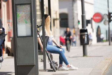 Casual caucasian teenager commuter with modern foldable urban electric scooter sitting on a bus stop bench waiting for metro city bus. Urban mobility concept.