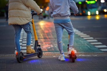 Couple rides around city at night on rent scooter in the evening. Trendy couple on electric scooter stand on crosswalk at night on city street. E-scooter or motorized vehicle for walking around.