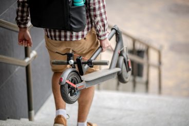 Young caucasian man in casual clothes with a backpack carries a folded electric scooter in his hand up the stairs of an office building. Man Carrying E-Scooter in folded position From Work.