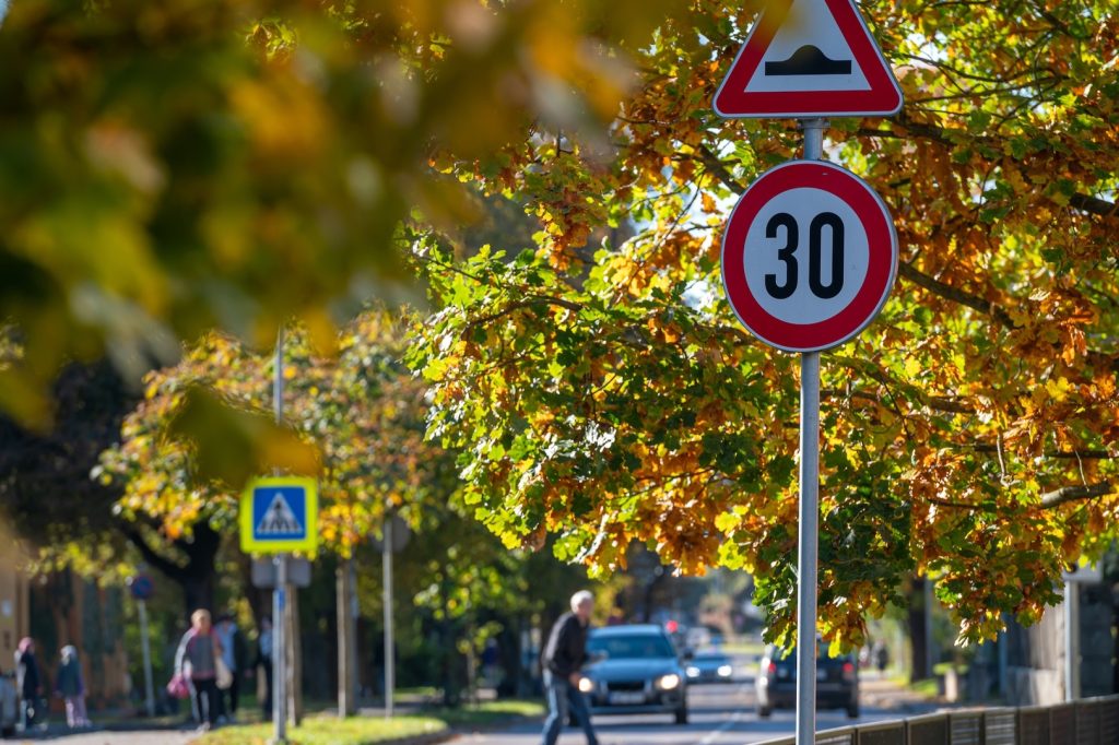 Speed limit signs beside a tree-lined street as pedestrians walk during autumn.